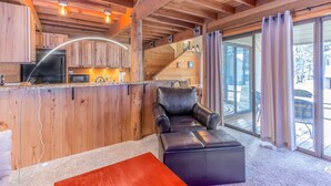Une cuisine moderne avec des armoires en bois et un réfrigérateur en acier inoxydable. Le salon est doté d’un canapé et d’un pouf en cuir noir, et une porte en verre donne accès à un patio extérieur.