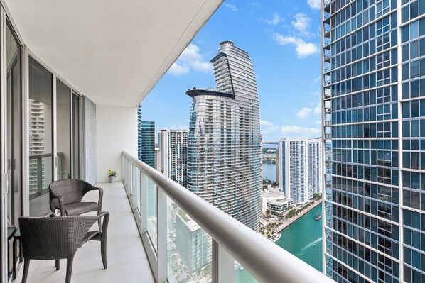 A balcony with a wicker chair, a view of a tall building, and a body of water.