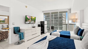 A hotel room with two beds, a TV, a desk, a chair, and a view of a cityscape through large windows. - 0142 Skyline Serenity 1B/1B (Miami)