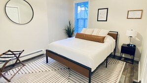 A bedroom with a bed, a ceiling fan, a mirror, a window, and a nightstand with a lamp.
