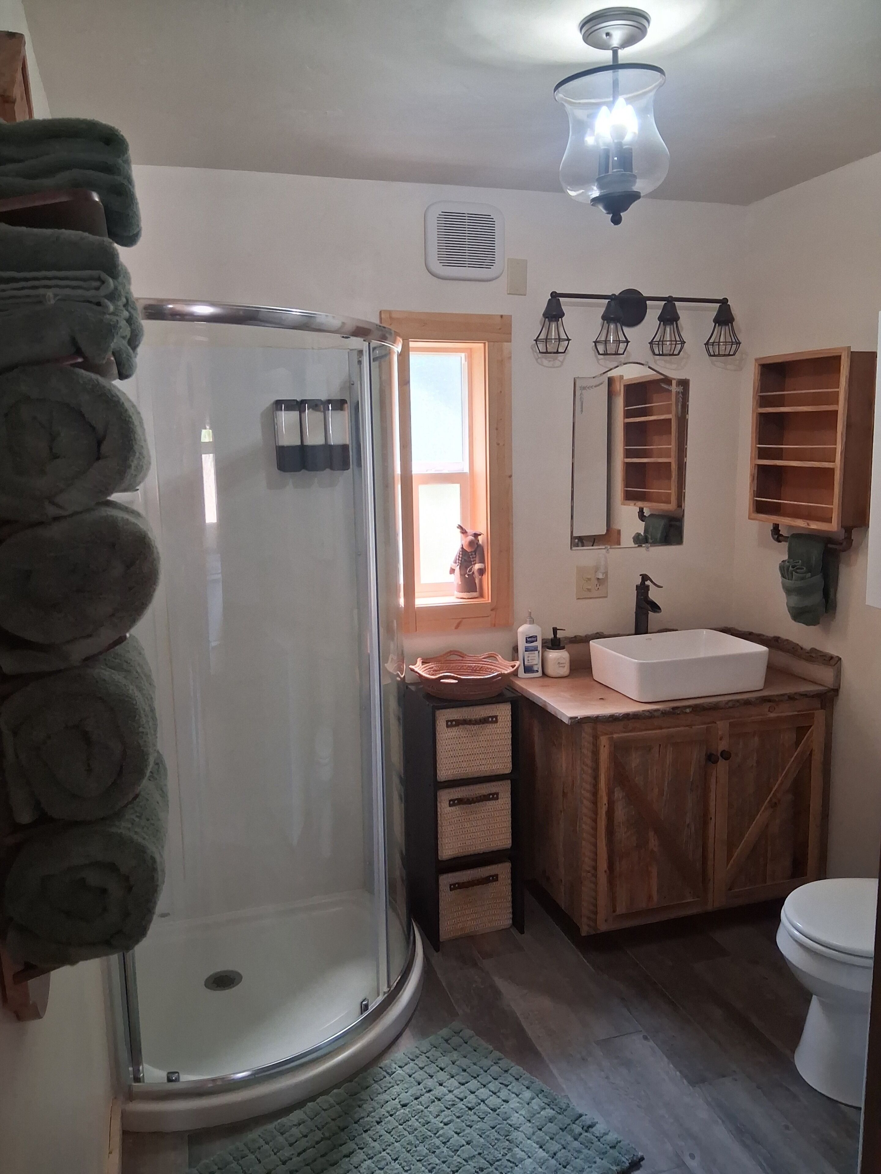 Un baño con cabina de ducha de vidrio, tocador de madera con lavamanos, inodoro y ventana con vistas.