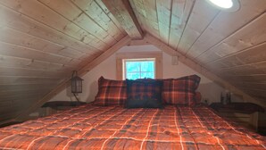 A loft bedroom with a sloped ceiling, a window, a bed with a plaid comforter, and a wall-mounted lamp.