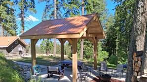 A wooden pavilion with a metal roof, surrounded by outdoor seating and a fire pit, set in a wooded area with a house in the background.