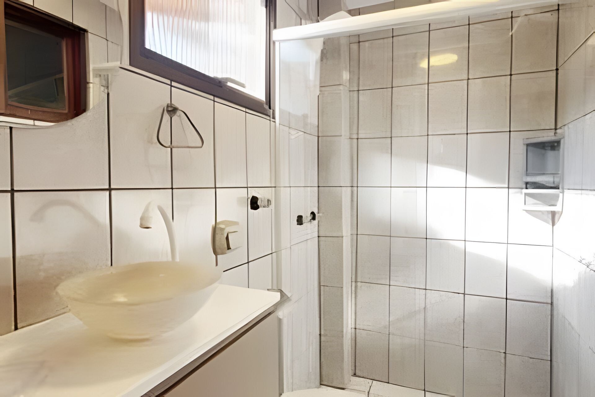 A bathroom with white tiles, a glass shower enclosure, a white sink with a glass bowl, and a window with blinds.