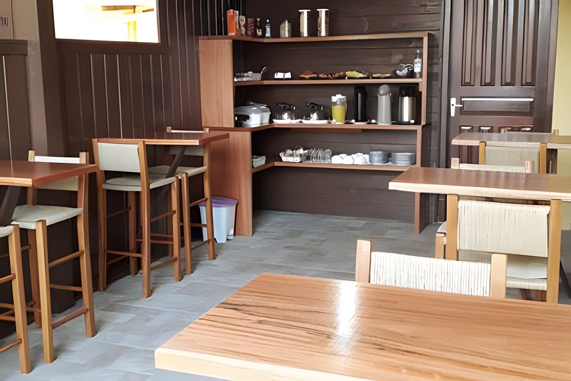 A restaurant with wooden tables and chairs, a kitchen area with shelves, and a trash bin.