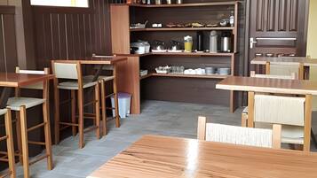 A restaurant with wooden tables and chairs, a kitchen area with shelves, and a trash bin.