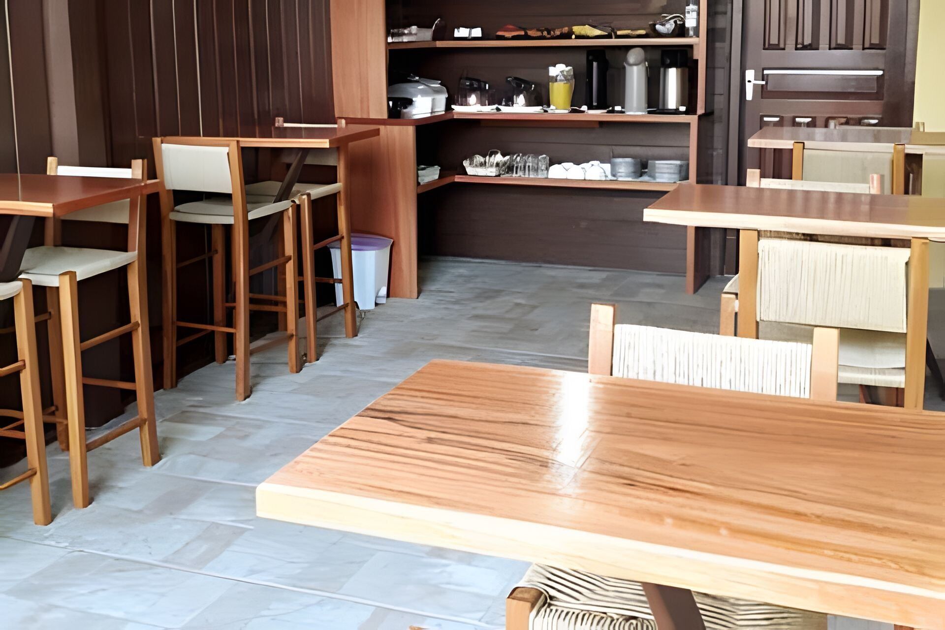 A restaurant with wooden tables and chairs, a tiled floor, and shelves with kitchen items.