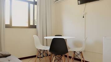 A small, well-lit room with a round table and four chairs, a flat-screen TV mounted on the wall, and a window with curtains.