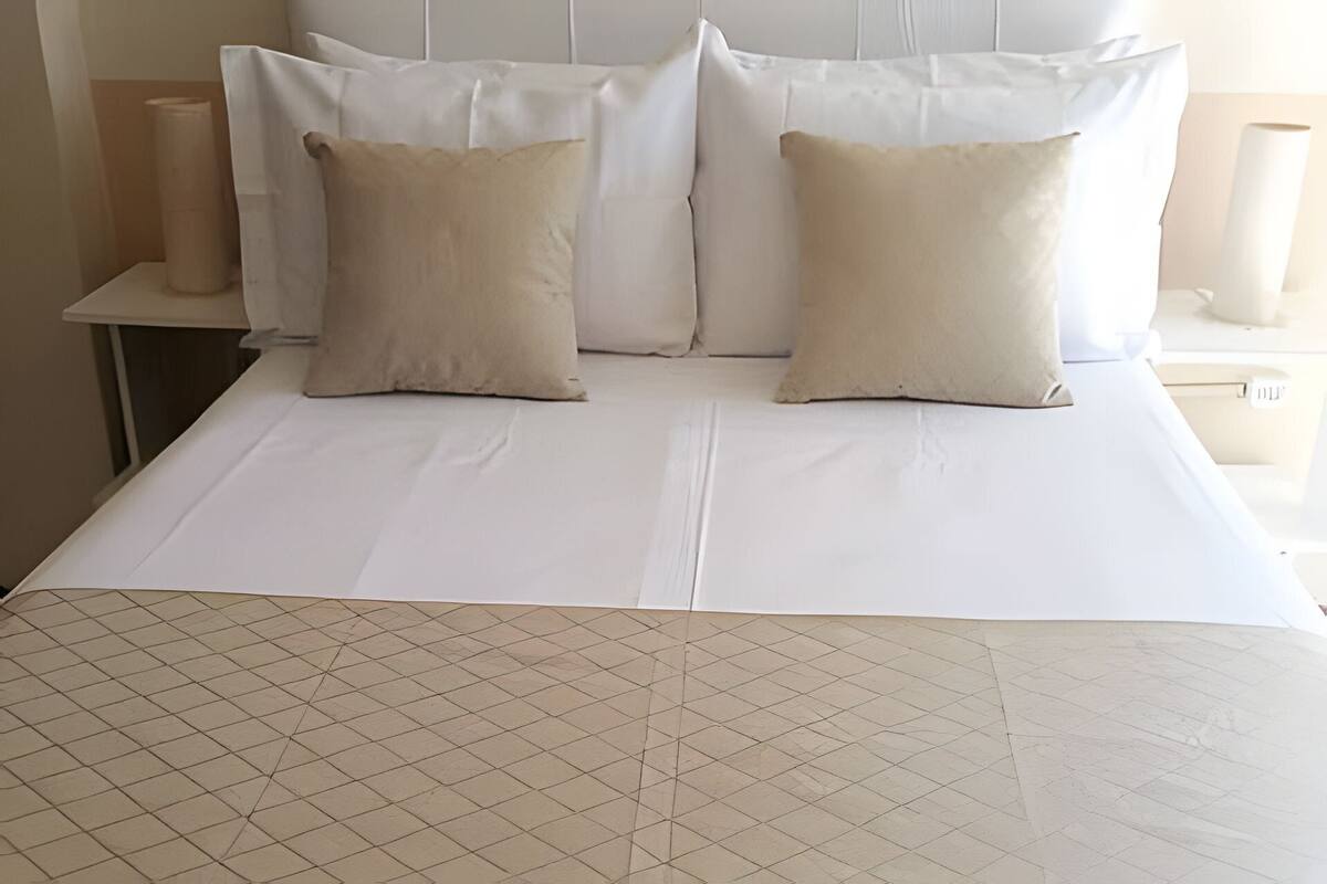 Una cama con colchón blanco y almohadas color beige, una pared lisa y una pequeña mesa con un rollo de toallas de papel.