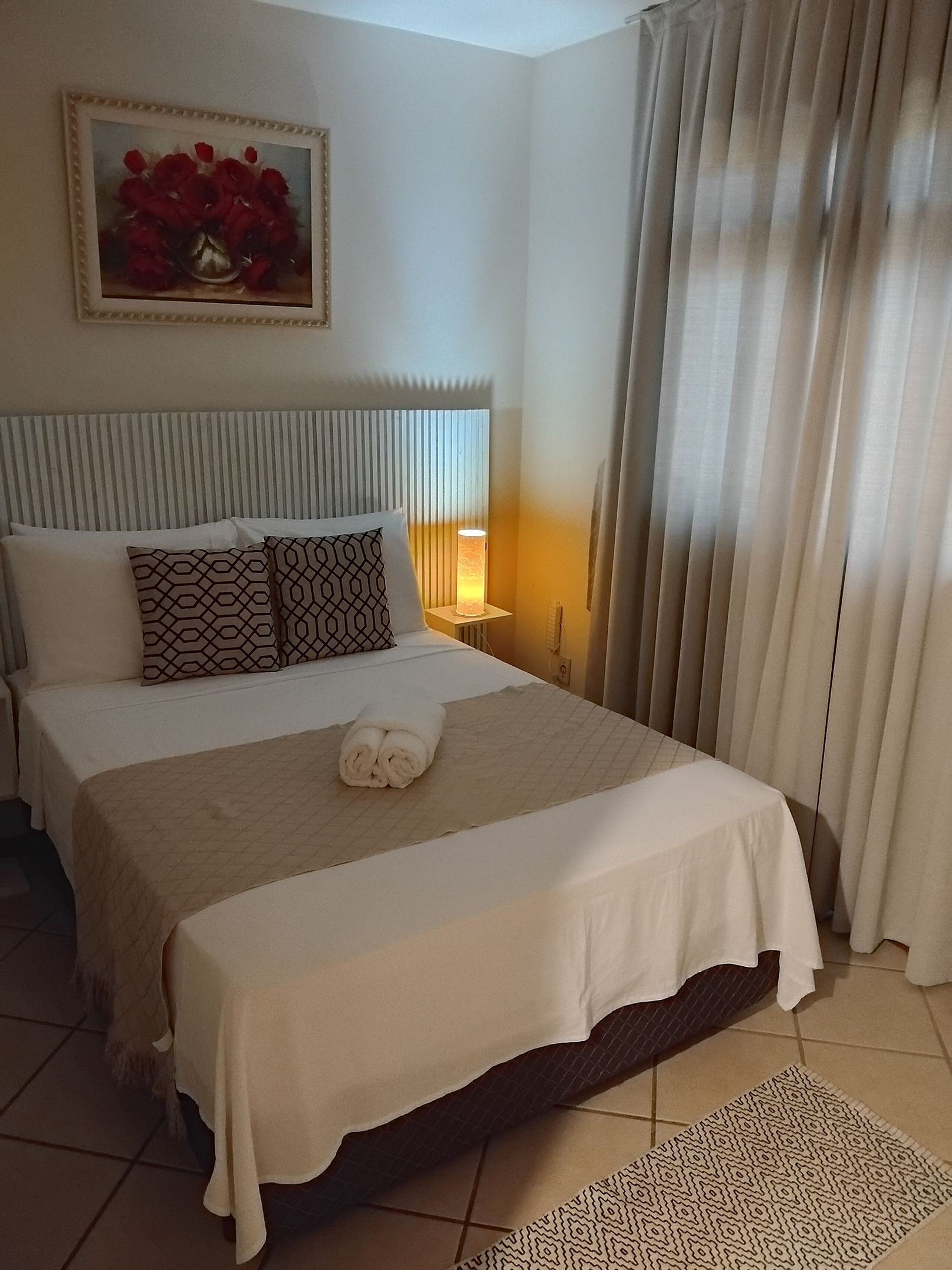 A neatly made bed with a white and patterned bedspread, a rolled towel, and two patterned pillows. There is a bedside lamp, a framed picture on the wall, and a window with curtains.