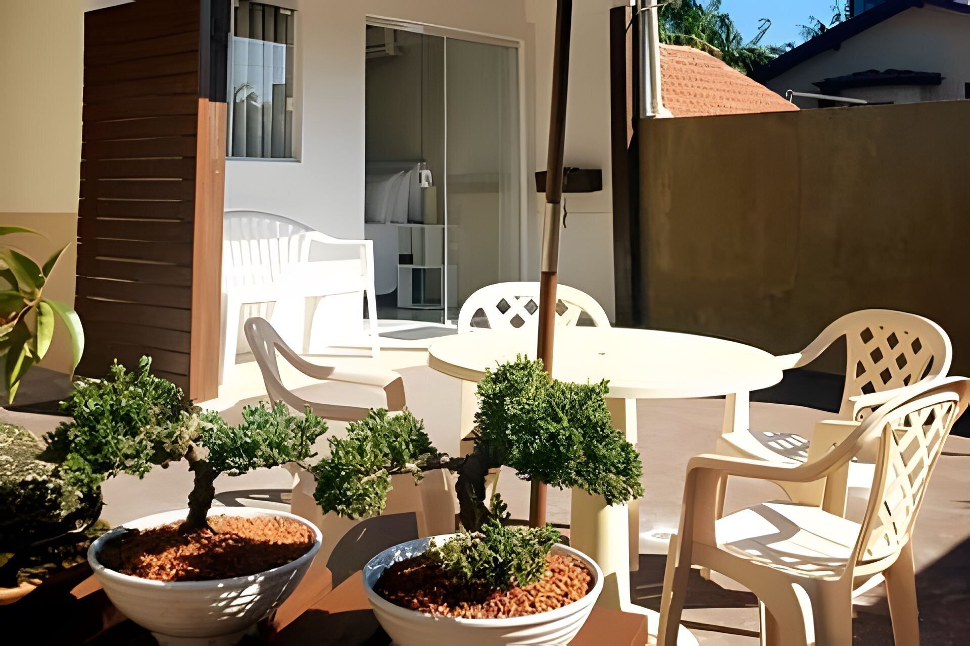 Un patio con una mesa redonda, dos sillas blancas y dos bonsáis en macetas.