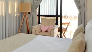 A hotel room with a large bed, a desk with a lamp, a chair, and a view of the outdoors through a window with curtains.