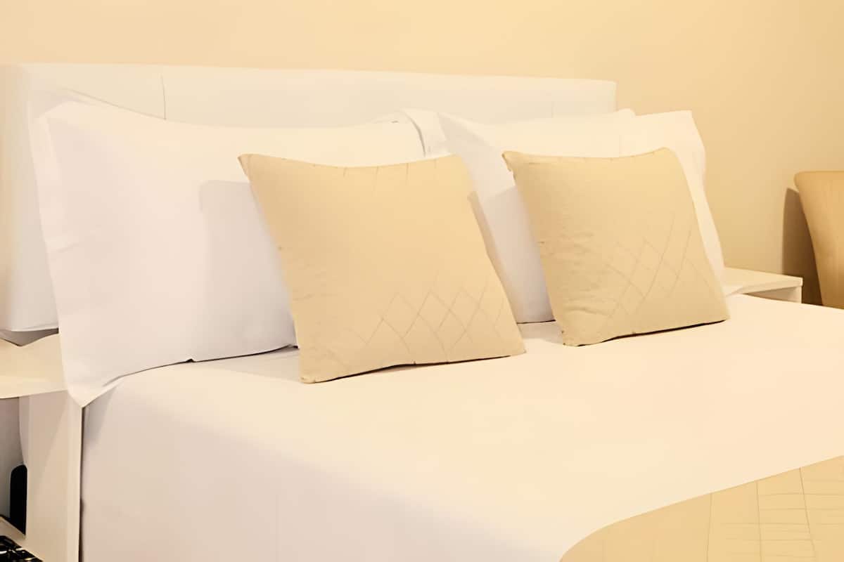 Una cama bien hecha con ropa de cama blanca y dos almohadas beige con diseño de rombos.