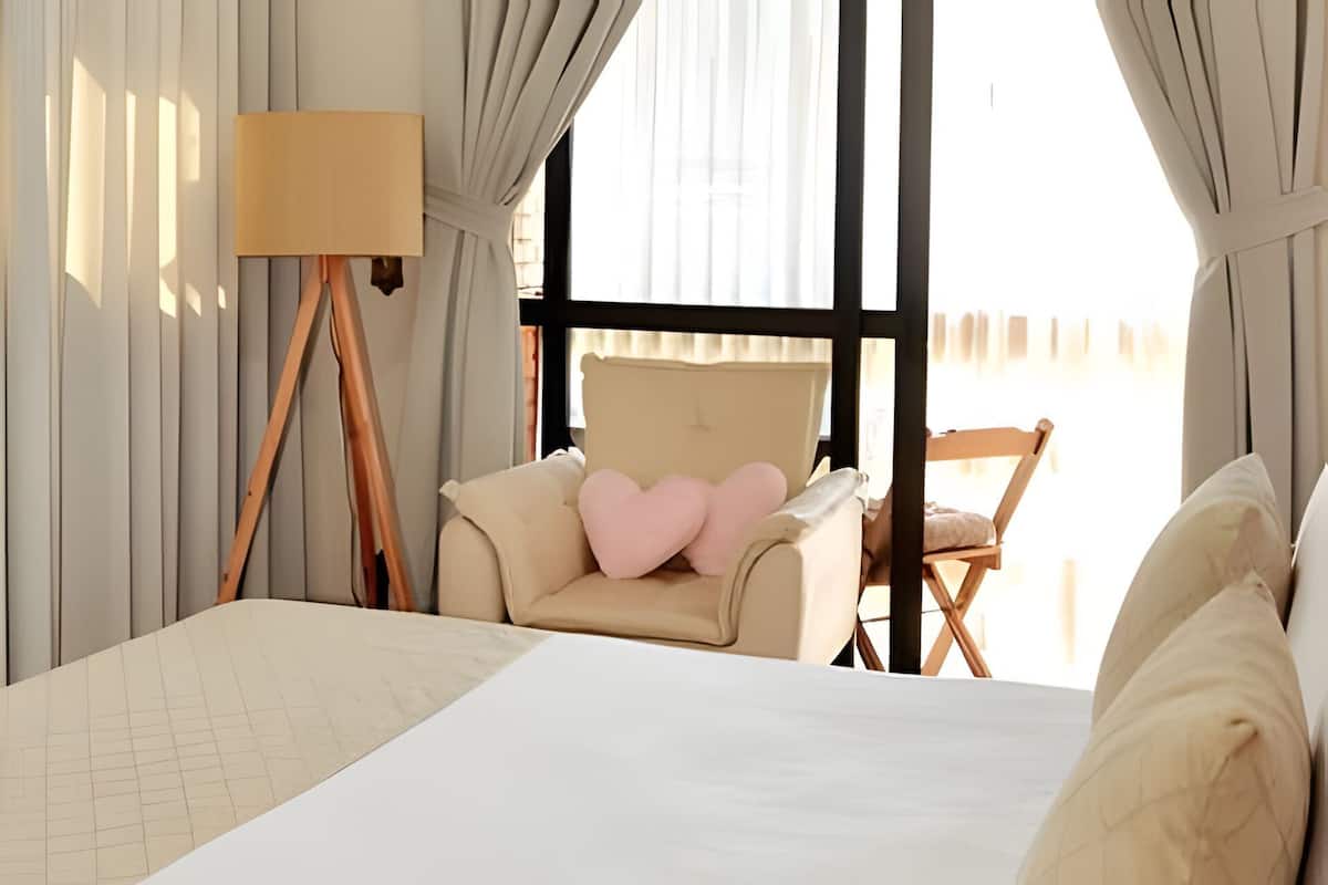Habitación de hotel con una cama grande, una silla con almohadas con forma de corazón, una lámpara de pie y vistas al exterior.