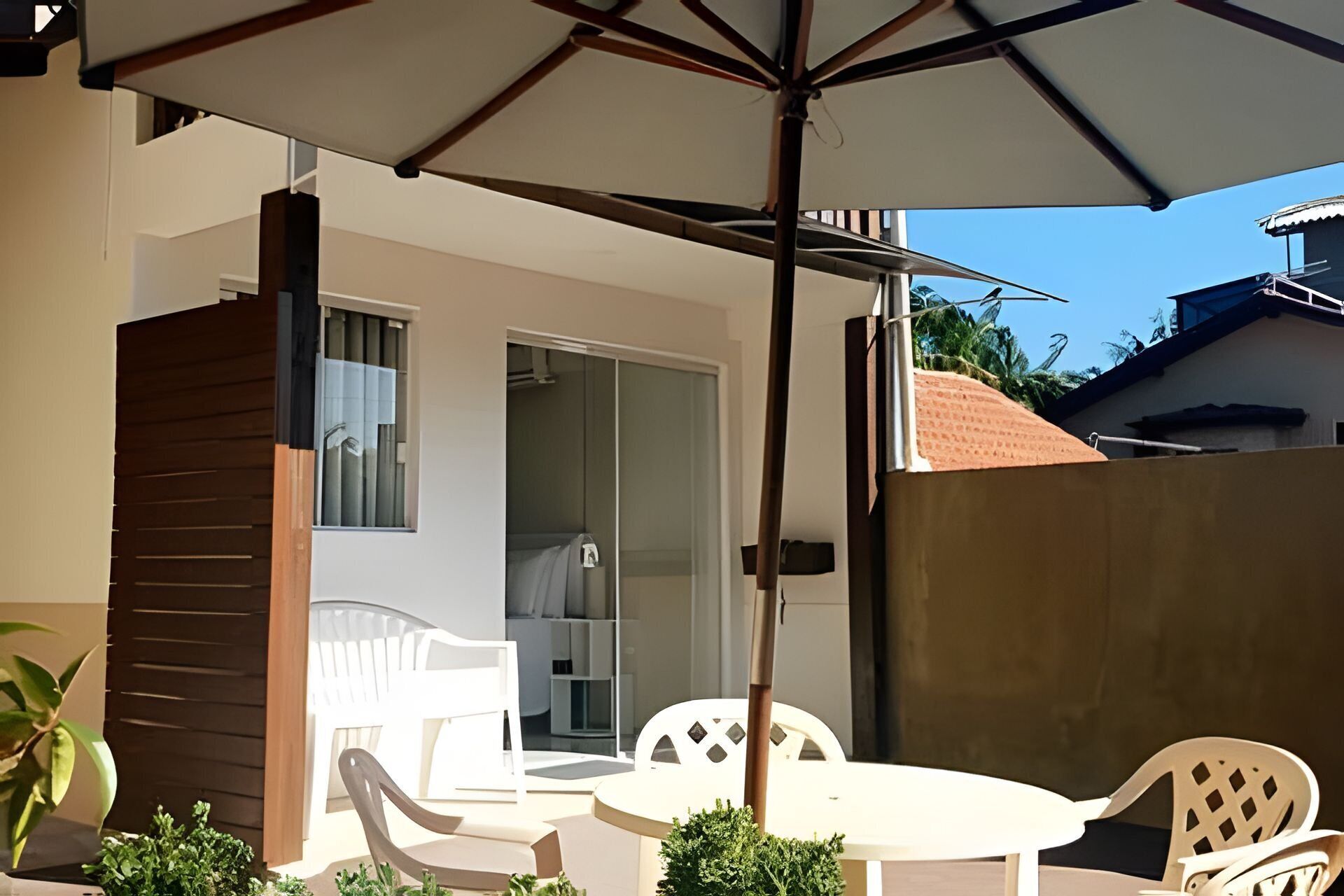 Un espace patio avec une table et des chaises blanches, un grand parasol et une vue sur l’intérieur du bâtiment.