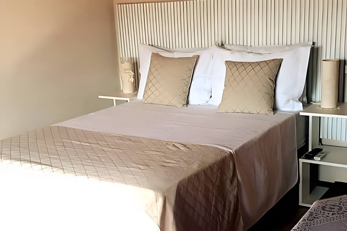 Una cama bien tendida con una colcha estampada, dos almohadas y una mesita de noche con una lámpara y un teléfono.