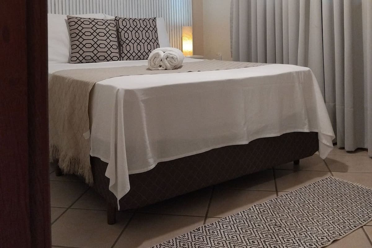 Una cama bien hecha con cubrecama blanco y dos almohadones estampados, un cuadro enmarcado con flores rojas en la pared y un ventanal con cortinas claras.