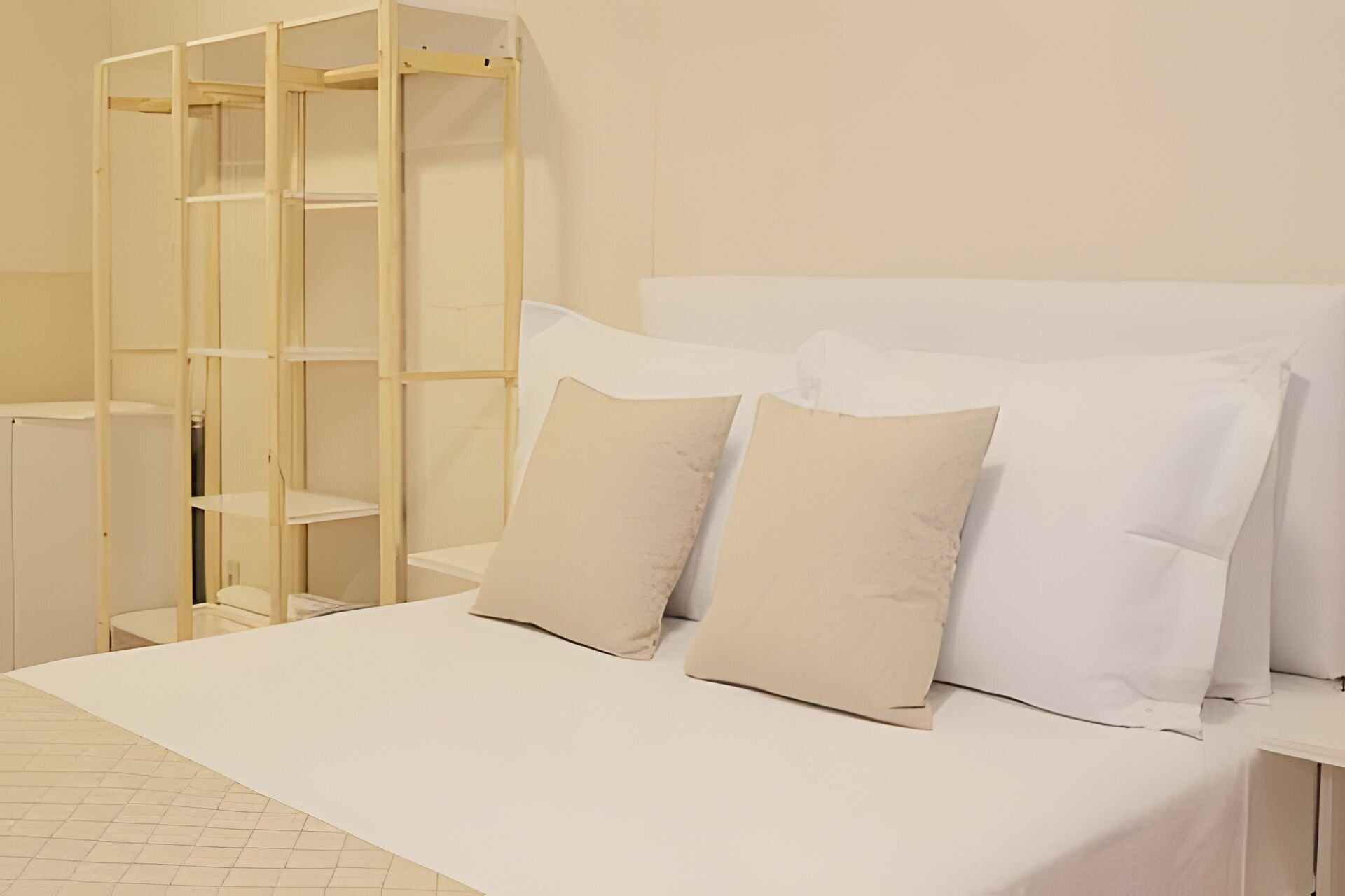 Una cama con ropa blanca y dos almohadas, una estantería de madera y una mesita de noche blanca.
