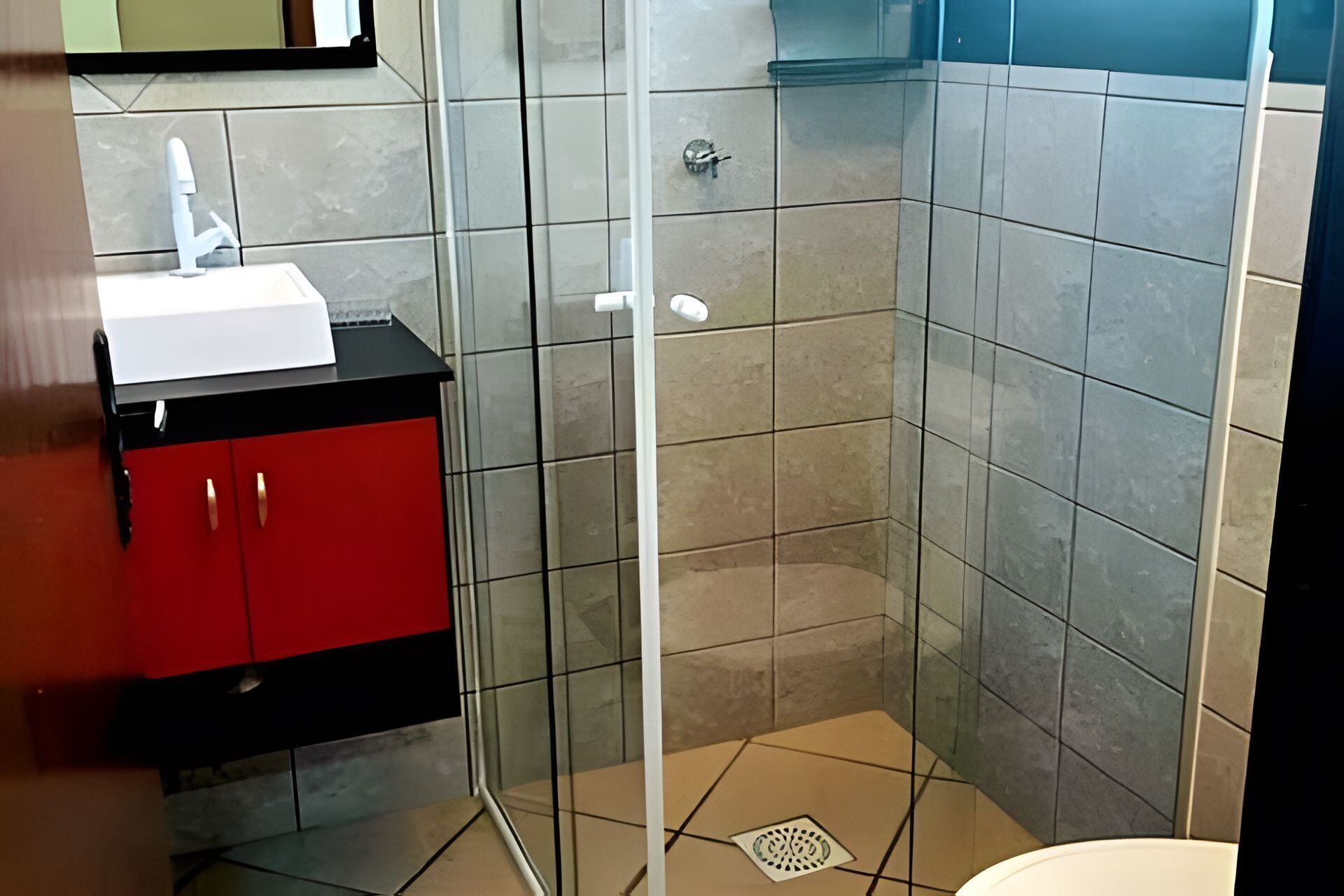 Un baño con cabina de ducha de vidrio, un mueble rojo con lavamanos blanco y paredes con azulejos.