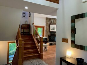 A staircase leading to a living area with a stone fireplace, wooden flooring, and a view of greenery outside.