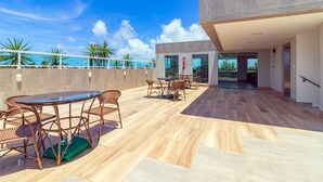 Una terraza en la azotea con zona de comedor, la entrada de un edificio moderno y vistas al océano.