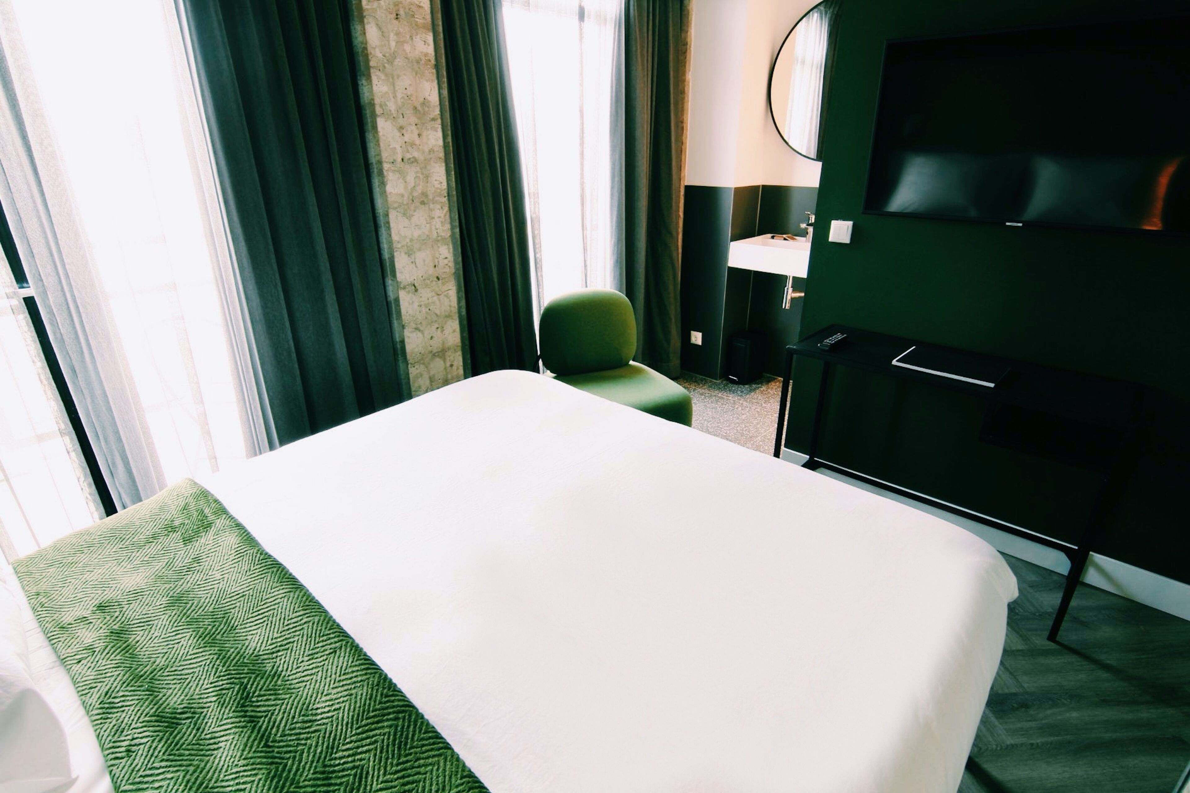 A hotel room with a bed, a green chair, a TV, and a desk with a lamp.