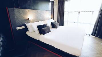 A modern hotel room with a large bed, dark headboard, and a view of the city through large windows.
