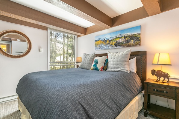 Une chambre avec un grand lit, une table de chevet avec une lampe, un tableau au mur et une fenêtre donnant sur des arbres.