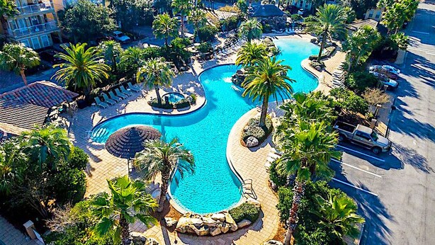 An aerial view of a resort with a large, winding pool surrounded by palm trees and lounge chairs. - Excellent Management Steps From Beach Pool (Destin)