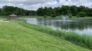 A gazebo near a pond with a fountain, surrounded by lush greenery and trees.