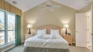 Une chambre à coucher avec un grand lit, deux tables de chevet avec des lampes, un ventilateur de plafond et une fenêtre avec des rideaux.
