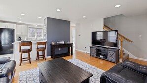 Una sala de estar moderna con una mesa de centro de madera oscura, un sofá de cuero negro y un televisor sobre un mueble de madera. Se observa un área de cocina con armarios blancos y un refrigerador negro al fondo.