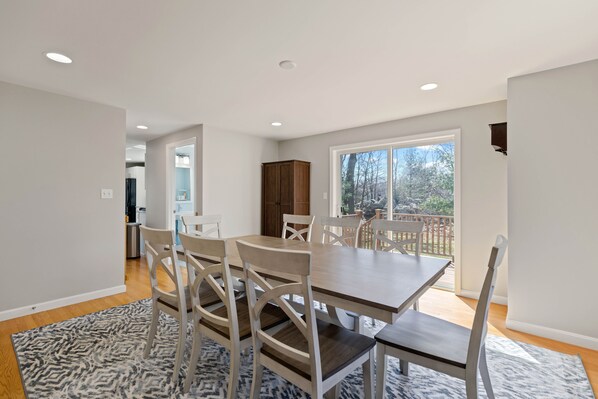 Comedor con una mesa grande y sillas, cocina al fondo y vistas al exterior.