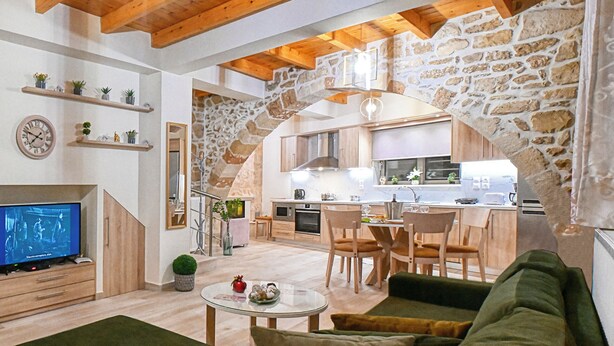 Een moderne woonkamer met een televisie, bank en eethoek. In de achtergrond is een keuken zichtbaar, met houten kasten en een stenen boog.
