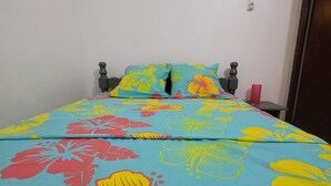 Ein Bett mit floralem Überwurf und zwei Kissen, ein Nachttisch mit einem roten Gegenstand sowie eine Tür rechts.