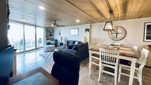 A spacious living area with a wooden ceiling, a dining table with chairs, a sofa, and a large window offering a view of the city. - "The Turtle Nest" a 3bed/2 bath Condo in St Regis (New Listing) (North Topsail Beach)