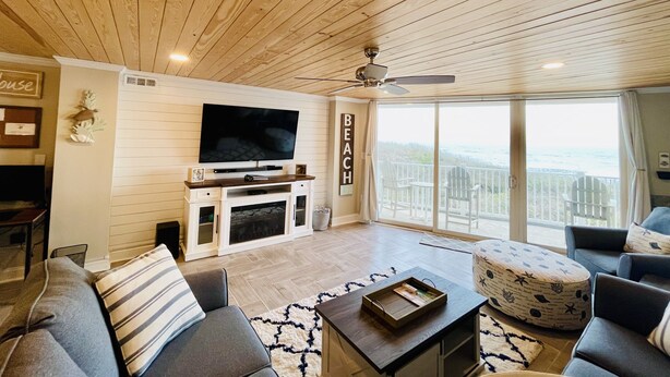 A spacious living room with a wooden ceiling, a flat-screen TV mounted on the wall, a fireplace, and a large window offering a view of the beach. - "The Turtle Nest" a 3bed/2 bath Condo in St Regis (New Listing) (North Topsail Beach)