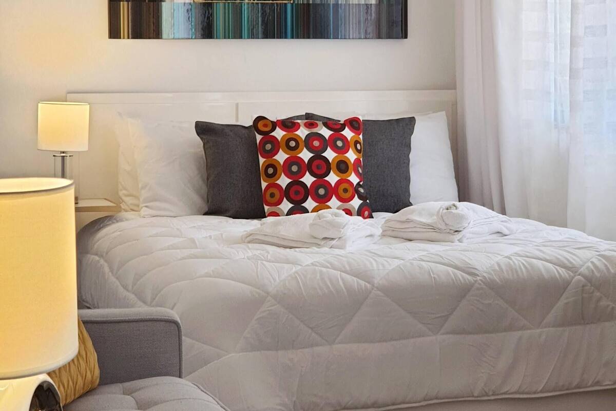 A neatly arranged bedroom with a bed, bedside table, lamp, and a modern abstract painting on the wall.