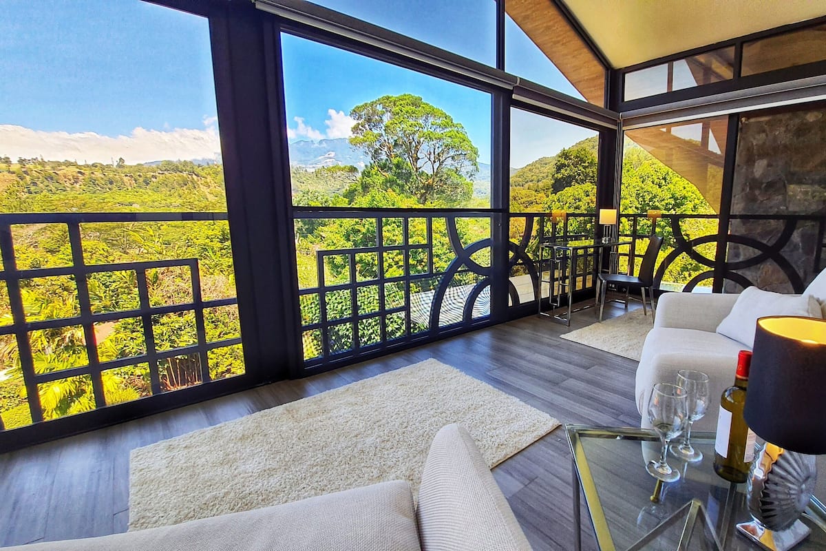 A modern room with large windows, a sofa, a glass table, and a view of a green landscape.