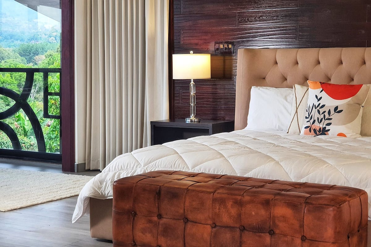 A bedroom with a large bed, a nightstand, a lamp, and a view of greenery through the window.
