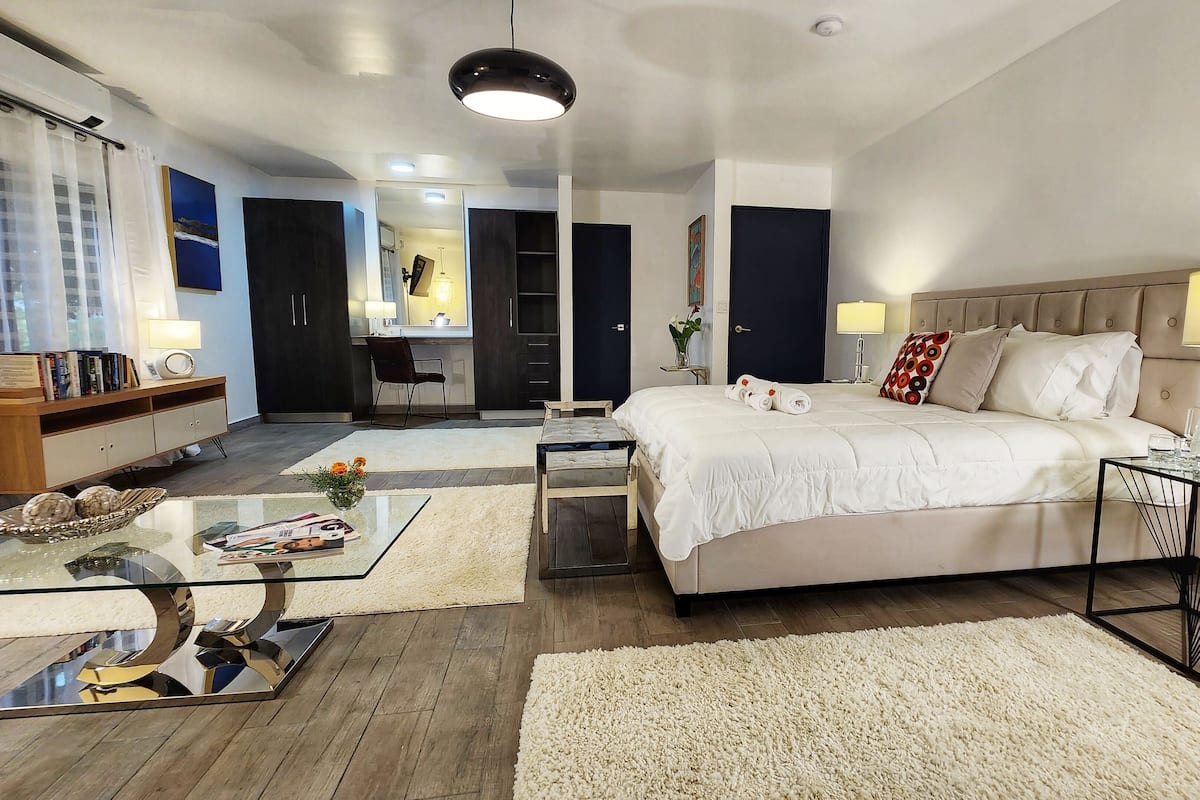 A modern hotel room with a large bed, a glass coffee table, a side table with a lamp, and a view of the bathroom and kitchen.