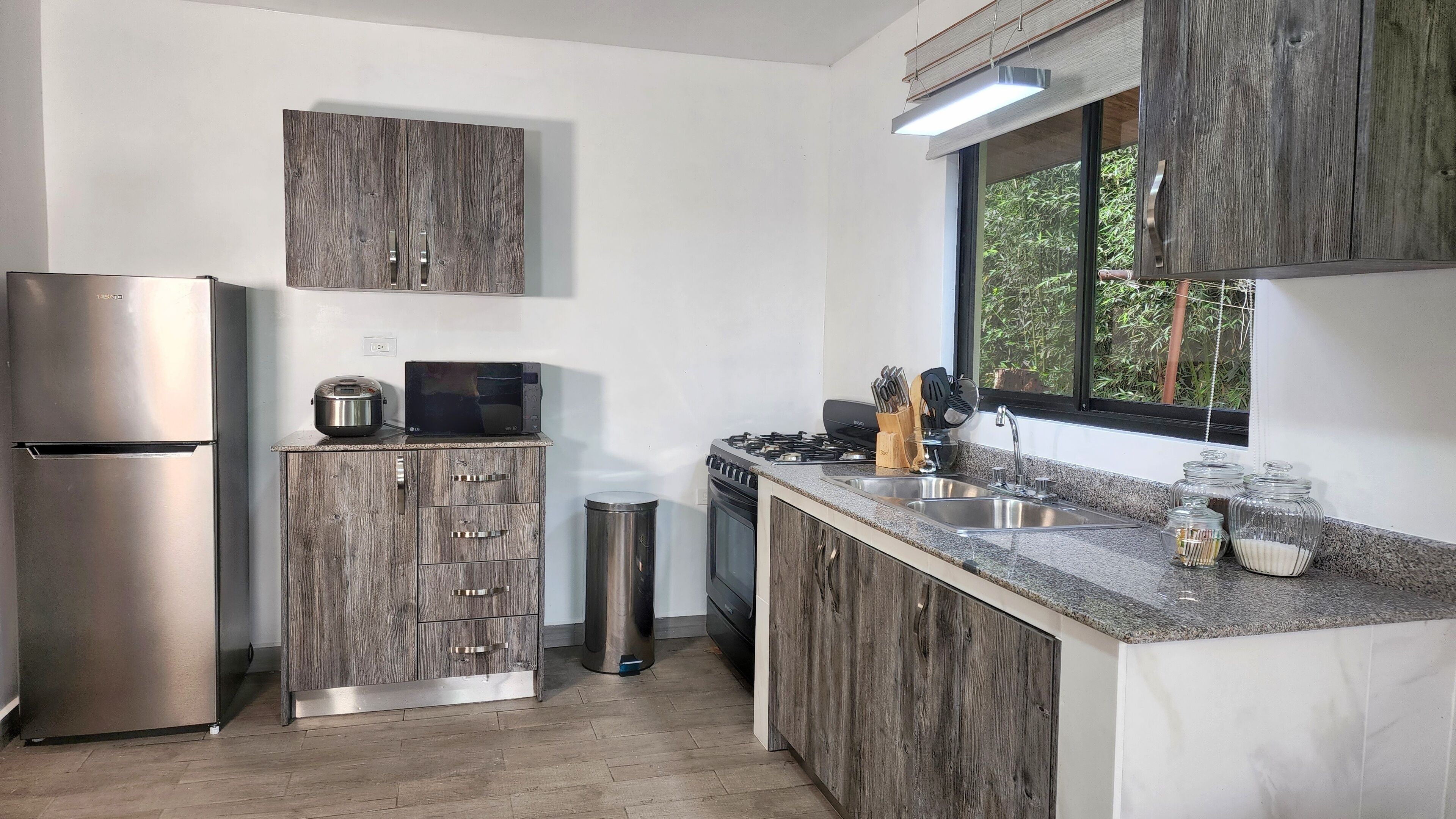 Una cocina moderna con un refrigerador de acero inoxidable, un microondas y una estufa a gas. Hay un gabinete de madera con cajones y un bote de basura. La cocina tiene una encimera de granito y una ventana con vista a zonas verdes.