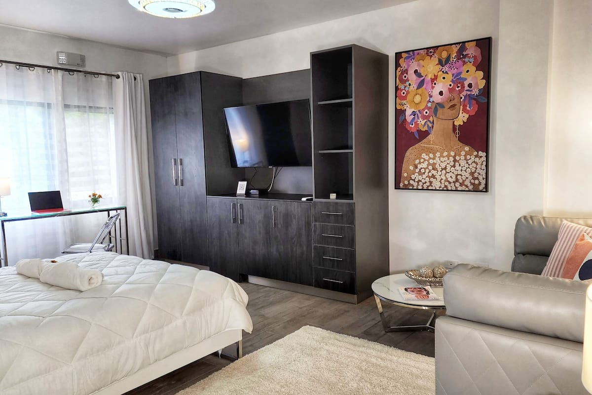 A modern hotel room with a bed, a sofa, a desk with a laptop, a TV, and a painting on the wall.