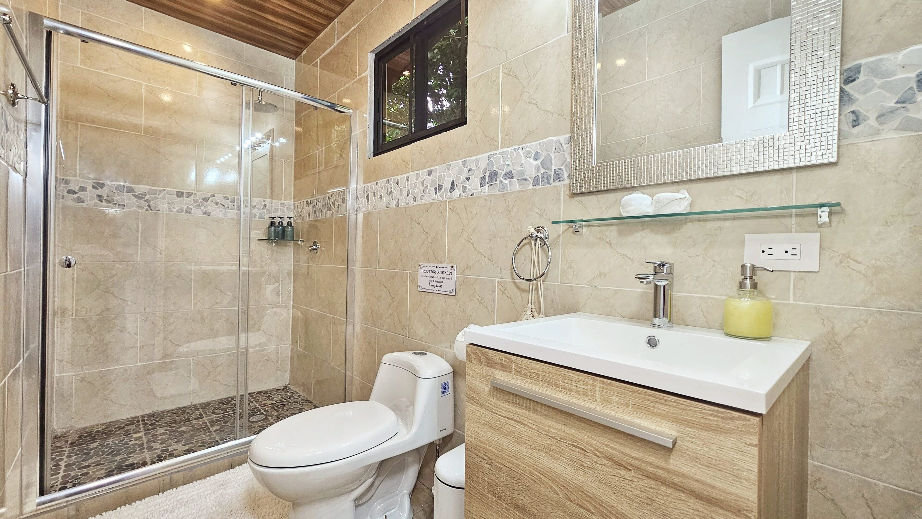 Un baño moderno con cabina de ducha de vidrio, inodoro, lavabo con espejo y un estante pequeño con artículos de tocador.
