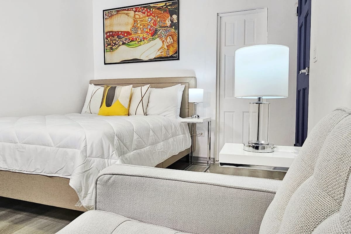 A neatly arranged hotel room with a bed, a bedside table with a lamp, a chair, and a painting on the wall.