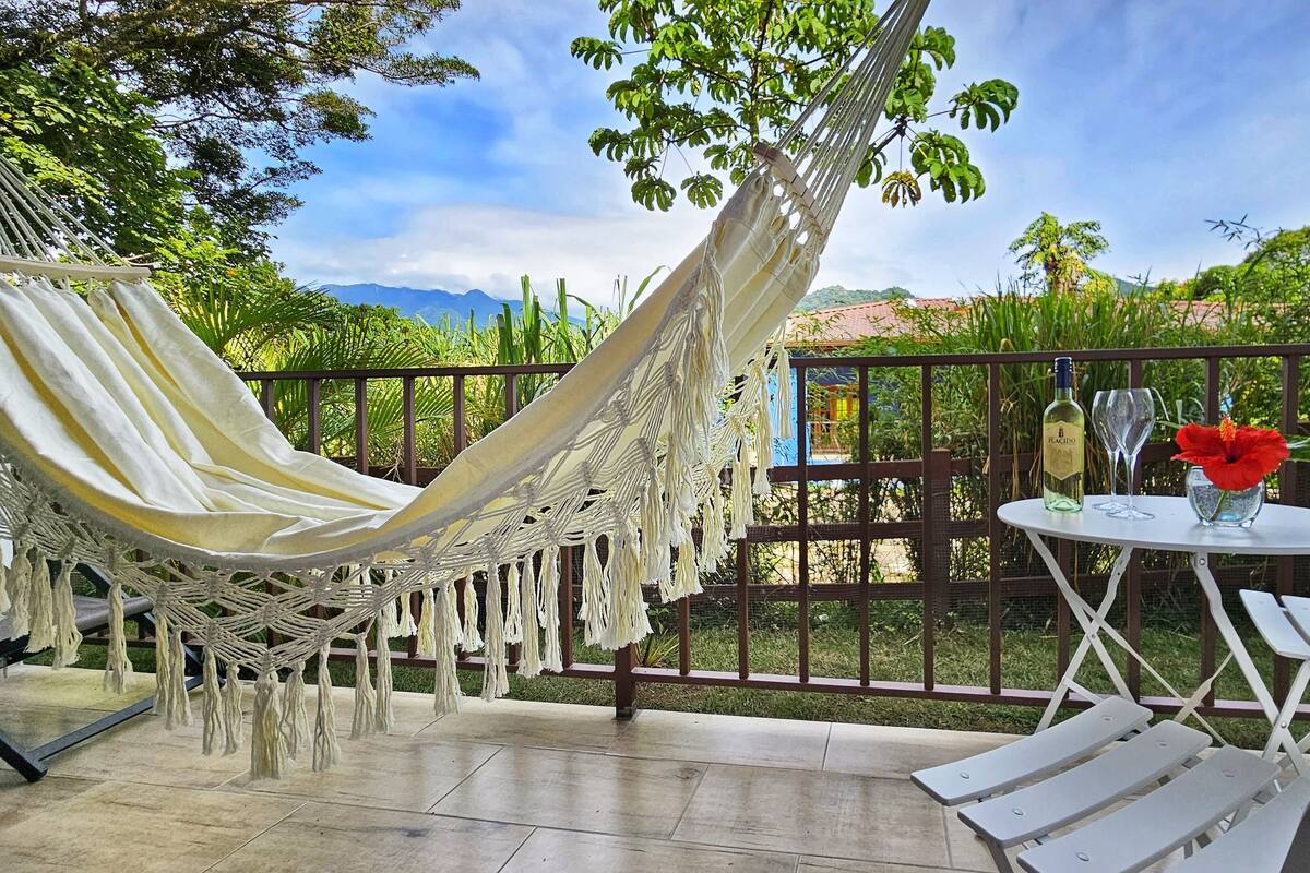 A hammock, a table with chairs, and a bottle of wine on a balcony with a view of greenery and mountains.