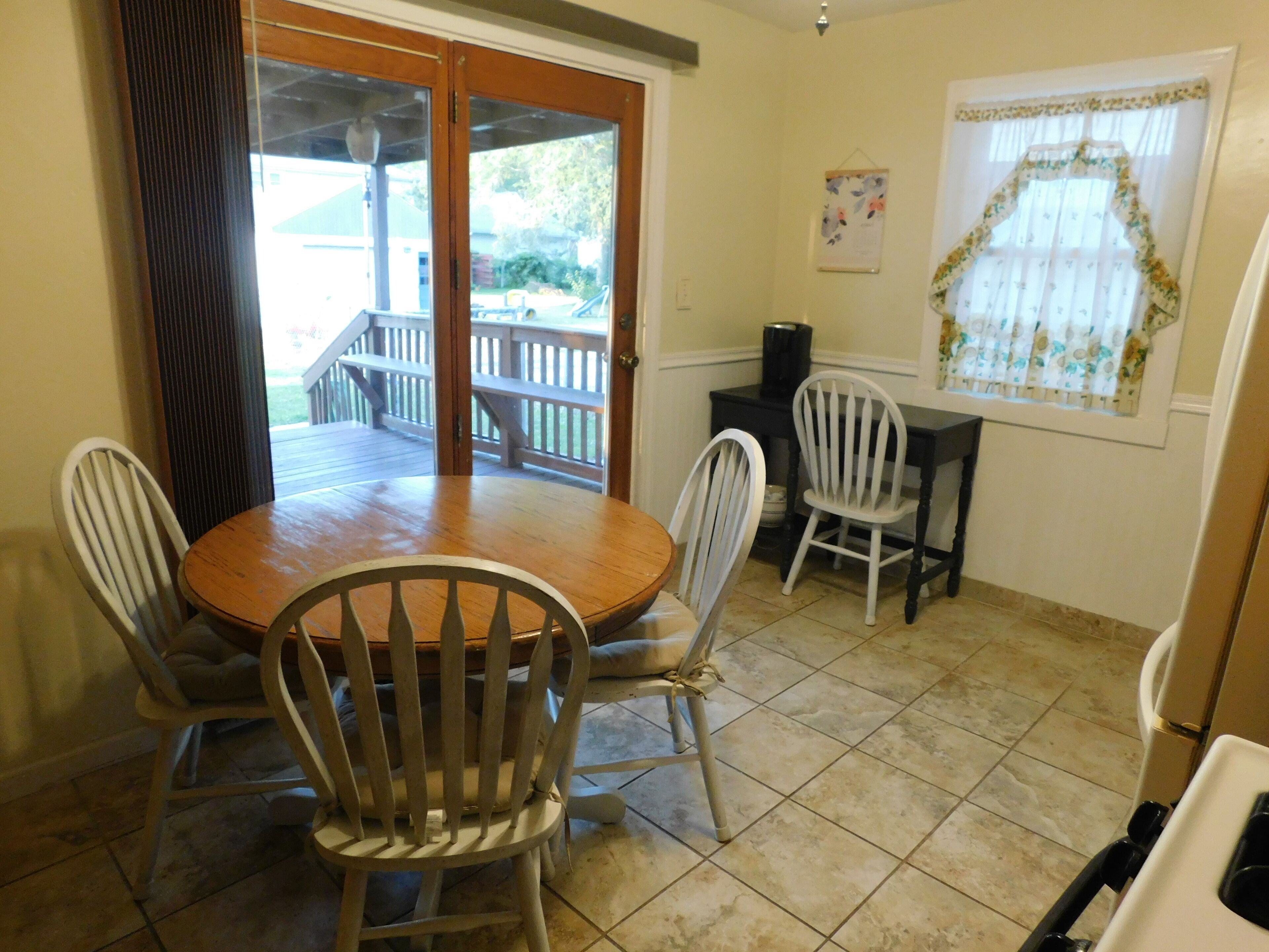 A dining area with a wooden table and four chairs, a black coffee machine, and a window with a floral curtain.