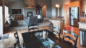 Uma sala de estar espaçosa com lareira, uma área de jantar com uma mesa comprida e uma cozinha com armários de madeira e uma geladeira de aço inoxidável.