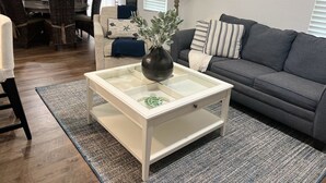 Ein modernes Wohnzimmer mit einem Couchtisch aus Glas, einem grauen Sofa und einem Essbereich im Hintergrund.
