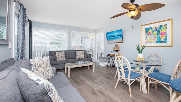 A modern living room with a grey sofa, a glass coffee table, wicker chairs, a ceiling fan, and a TV on the wall. - Spacious 2 bd/2b Arcadian Dunes near Shore Drive, Ocean Annie's, golf & beach (Myrtle Beach)